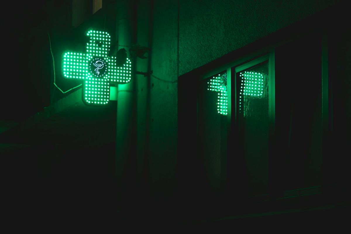 A green pharmacy cross glowing on a dark street at night, reflected in a window