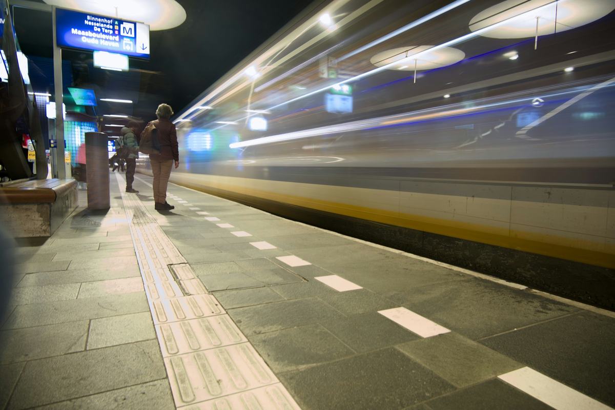 A traveller with a backpack stands alone on a metro platform at night as a train pulls away in a blur