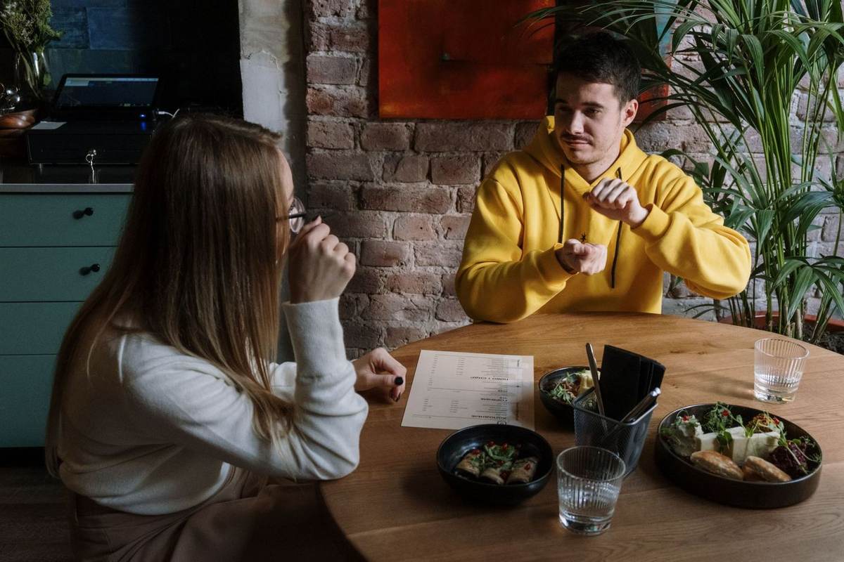 An einem Restauranttisch unterhält sich ein Mann in einem gelben Kapuzenpulli in Gebärdensprache mit der Frau ihm gegenüber; zwischen ihnen liegen Teller und eine Speisekarte